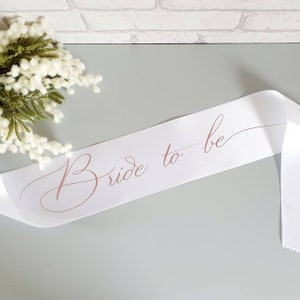 May include: White satin sash with the words "Bride to be" written in cursive in a rose gold color.