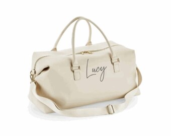 May include: A cream-colored canvas duffel bag with a gold zipper and a black leather strap. The bag has the name "Lucy" written on it in a cursive font.