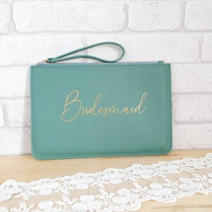 May include: A teal green clutch purse with a gold zipper and a gold handle. The purse has the word "Bridemaid" written in gold script on the front.