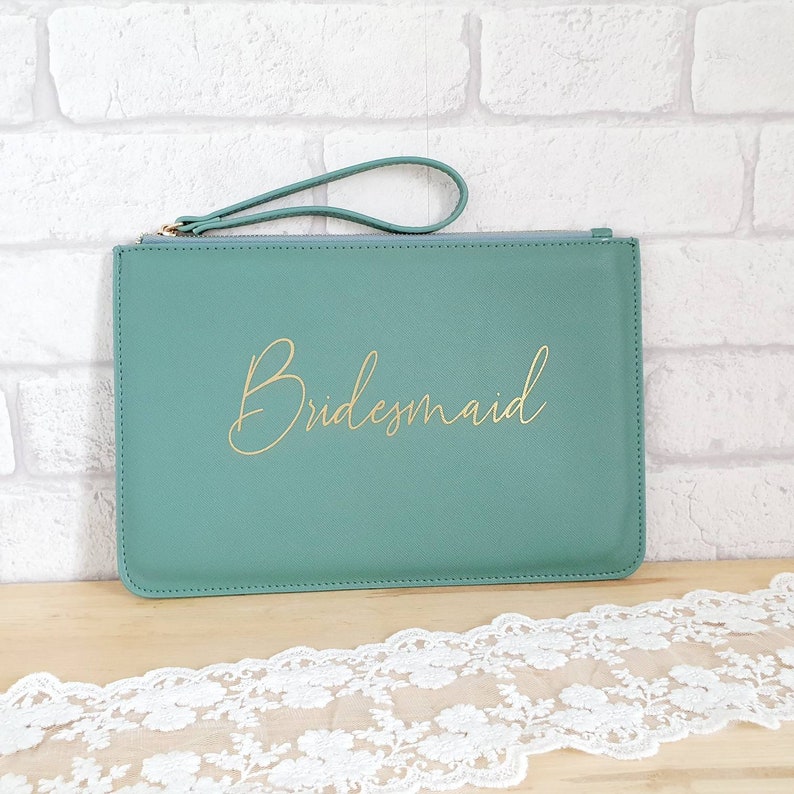 May include: A teal green clutch purse with a gold zipper and a gold handle. The purse has the word "Bridemaid" written in gold script on the front.