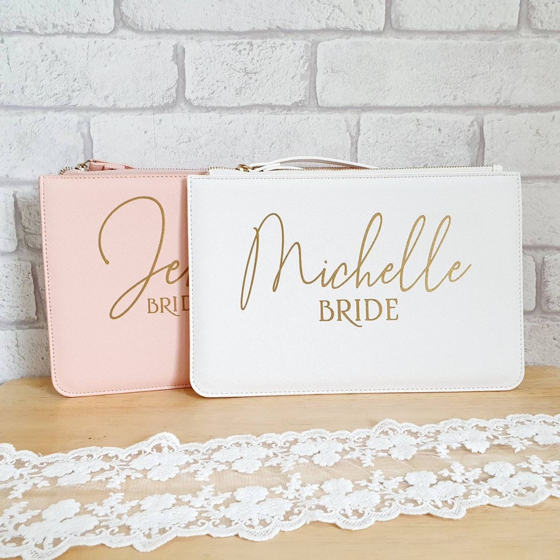 May include: Two white zippered clutches with gold lettering. The clutch on the left is pink and says "Jess Bride". The clutch on the right is white and says "Michelle Bride".