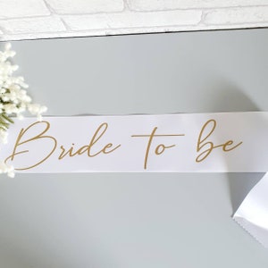 Puede incluir: Banda de satén blanco con las palabras "Bride to Be" escritas en letras doradas.