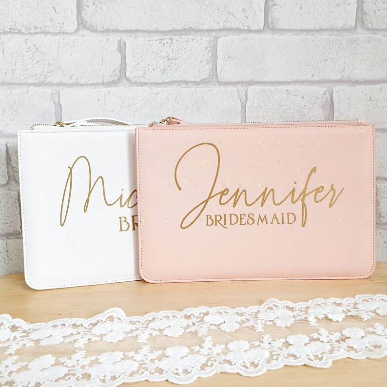 May include: Two personalized cosmetic bags, one white with the name "Mic" and the title "Bride" and one pink with the name "Jennifer" and the title "Bridesmaid".