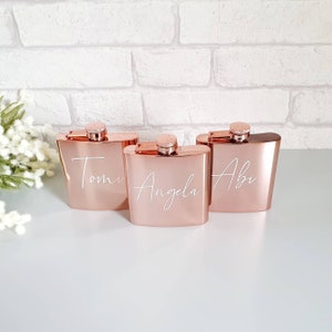 May include: Three rose gold flasks with screw-on caps. Each flask has a name in white script: Tomi, Angela, and Abi. The flasks are rectangular with rounded corners and sit on a white surface. The background is a white brick wall.