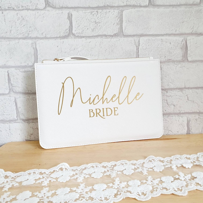 May include: White clutch bag with a gold zipper and the text "Michelle Bride" in gold script.