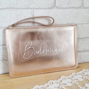 May include: A rose gold clutch purse with a white leather strap and the word "Bridemaid" written in white script.