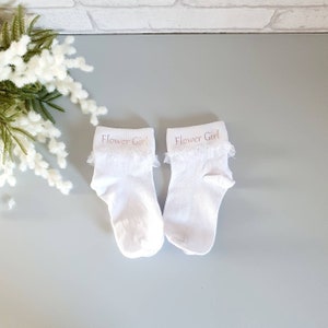 Puede incluir: Un par de calcetines blancos de tobillo con ribete de encaje y las palabras "Flower Girl" bordadas en rosa en la parte superior de cada calcet&iacute;n.