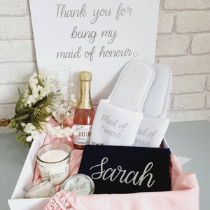 Puede incluir: Una caja de regalo blanca llena de artículos para una dama de honor. La caja incluye una botella de rosado espumoso, dos pantuflas blancas, una vela, una bolsa negra con el nombre "Sarah" bordado y un borde de encaje rosa.