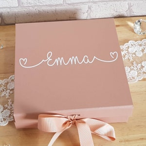 Puede incluir: Una caja de regalo rosa claro con una cinta blanca y el nombre "Emma" escrito en cursiva con corazones a cada lado.