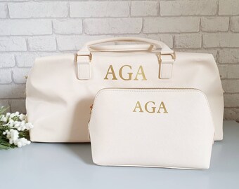 May include: A set of two cream-colored travel bags with gold zippers and the initials "AGA" embroidered in gold on the front of each bag.