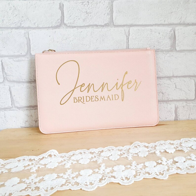 May include: A pale pink zippered pouch with the name "Jennifer" and the word "BRIDESMAID" written in gold lettering.