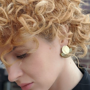 Puede incluir: Una mujer con el pelo rubio rizado que lleva una camisa negra y pendientes geom&eacute;tricos dorados y verdes.