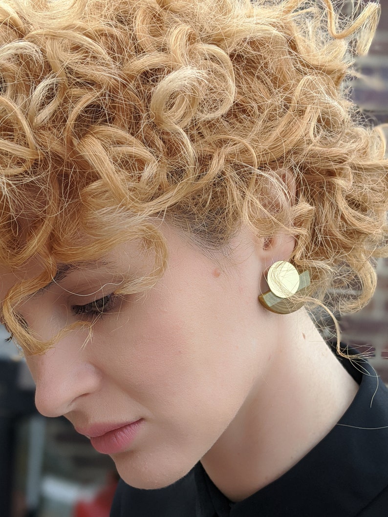 Puede incluir: Una mujer con el pelo rubio rizado que lleva una camisa negra y pendientes geom&eacute;tricos dorados y verdes.
