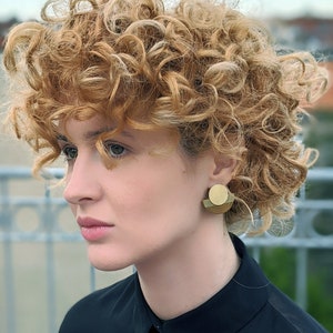 Puede incluir: Una mujer con el pelo rizado casta&ntilde;o claro lleva una camisa negra con cuello y pendientes geom&eacute;tricos dorados.