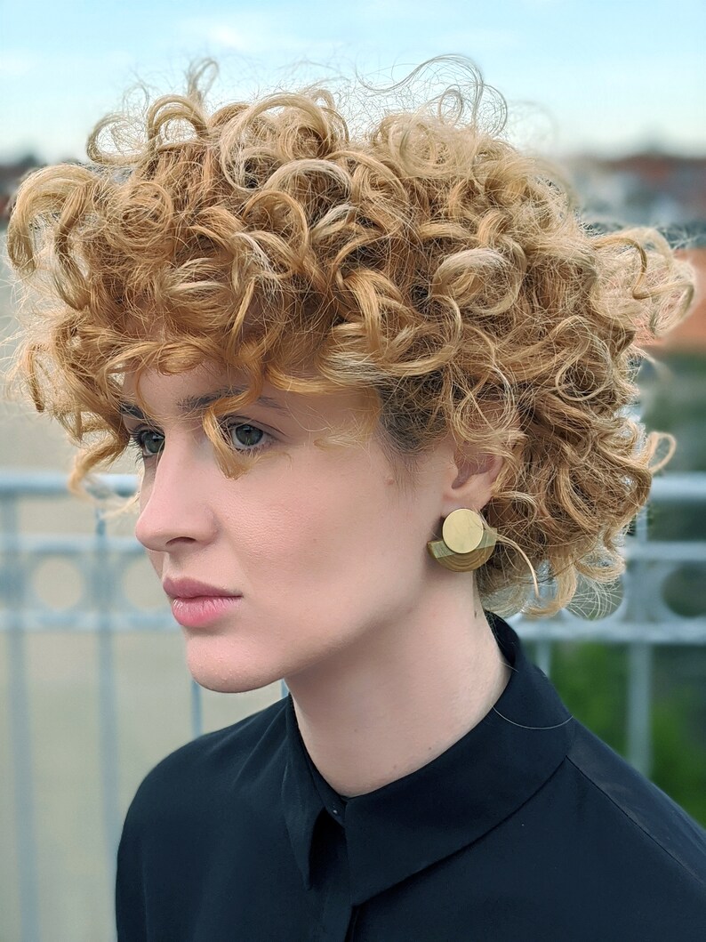 Puede incluir: Una mujer con el pelo rizado casta&ntilde;o claro lleva una camisa negra con cuello y pendientes geom&eacute;tricos dorados.