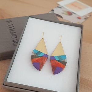 Puede incluir: Un par de pendientes geom&eacute;tricos coloridos con detalles dorados. Los pendientes est&aacute;n en una caja de regalo con el texto "WOODLANDarie" impreso en la caja.