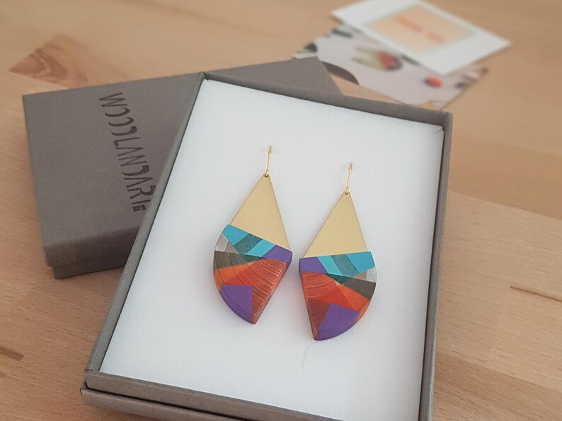 Puede incluir: Un par de pendientes geom&eacute;tricos coloridos con detalles dorados. Los pendientes est&aacute;n en una caja de regalo con el texto "WOODLANDarie" impreso en la caja.