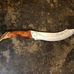 May include: A silver-colored dagger with a brown wooden handle and gold accents. The blade has a curved shape and is inscribed with a decorative design. The inscription reads "Aiya Eärendil Elenion Ancalima!"