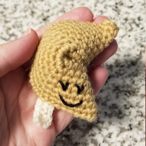 May include: A crocheted fortune cookie with a smiling face. The cookie is beige with white stitching and black embroidery.