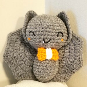 Puede incluir: Un murciélago de crochet gris con una pajarita naranja y blanca. El murciélago tiene una cara sonriente con ojos negros y una boca cosida.
