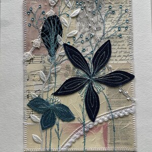 May include: A mixed media textile art piece featuring layered paper with printed text and musical notation, embellished with dark blue fabric flower appliqués, white leaf cutouts, teal embroidery, and small blue rhinestones, finished with a zigzag stitched border.