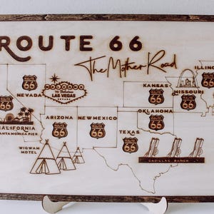 Puede incluir: Un letrero de madera con un mapa de la Ruta 66, con el texto "Route 66 The Mother Road" y varios puntos de referencia a lo largo de la ruta, incluido el Wigwam Motel, Cadillac Ranch y el muelle de Santa M&oacute;nica.