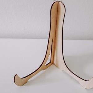 Puede incluir: Un caballete de madera con un acabado natural. El caballete tiene tres patas y est&aacute; dise&ntilde;ado para sostener un objeto peque&ntilde;o, como un marco de fotos o un plato.