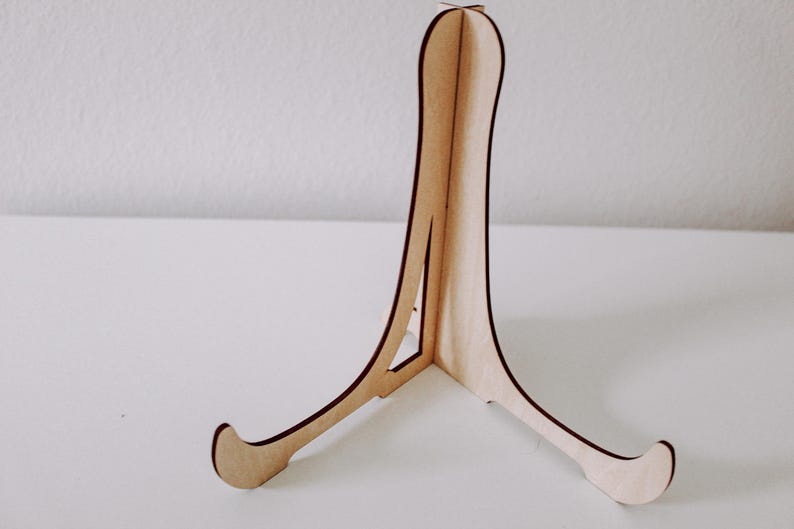 Puede incluir: Un caballete de madera con un acabado natural. El caballete tiene tres patas y est&aacute; dise&ntilde;ado para sostener un objeto peque&ntilde;o, como un marco de fotos o un plato.