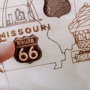 Puede incluir: Un mapa de madera de los Estados Unidos con un dedo apuntando a la Ruta 66 en Misuri. El mapa tambi&eacute;n incluye un dibujo del Arco Gateway en St. Louis y un restaurante vintage.