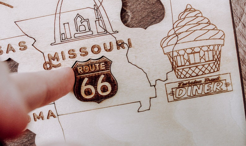 Puede incluir: Un mapa de madera de los Estados Unidos con un dedo apuntando a la Ruta 66 en Misuri. El mapa tambi&eacute;n incluye un dibujo del Arco Gateway en St. Louis y un restaurante vintage.