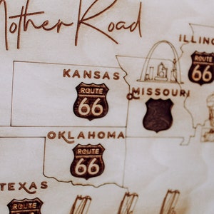 Puede incluir: Un mapa de madera de la Ruta 66, que muestra los estados de Illinois, Misuri, Kansas, Oklahoma y Texas. El mapa presenta el escudo ic&oacute;nico de la Ruta 66 con el n&uacute;mero 66 en cada estado. El mapa es de color marr&oacute;n claro con l&iacute;neas y texto negros.