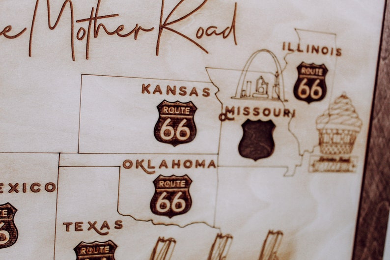 Puede incluir: Un mapa de madera de la Ruta 66, que muestra los estados de Illinois, Misuri, Kansas, Oklahoma y Texas. El mapa presenta el escudo ic&oacute;nico de la Ruta 66 con el n&uacute;mero 66 en cada estado. El mapa es de color marr&oacute;n claro con l&iacute;neas y texto negros.