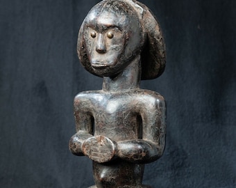 Puede incluir: Una escultura de madera tallada a mano en color marrón oscuro que representa una figura de pie con cabeza redondeada, orejas prominentes y superficie texturizada. Las manos están situadas frente al torso, sosteniendo un objeto pequeño y plano.