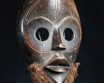 May include: A wooden mask with a human face, carved with detailed features. The mask has large, round eyes, a prominent nose, and a wide mouth. The mask is painted in shades of brown and black, and has a fringe of brown fibers around the bottom.