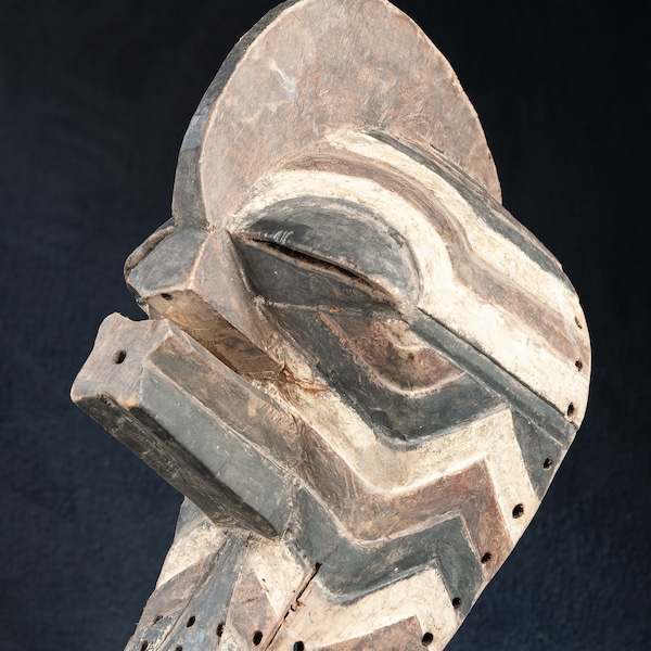 May include: A wooden mask with a geometric design in black, white, and brown. The mask has a large, rounded top and a long, narrow snout. It is carved with intricate details and has a number of small holes.