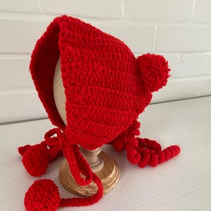 May include: A bright red, crocheted bonnet with a rounded crown and a pair of long ties with pom-poms. The bonnet is displayed on a wooden stand against a white brick wall. The bonnet has a textured appearance.