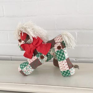 May include: A stuffed horse toy crafted from patchwork fabric featuring red, green, and white patterns. The horse has a white yarn mane and tail, a red polka-dot fabric bow at the neck, and brown button accents at the joints and eyes.