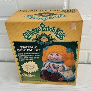 May include: A vintage Cabbage Patch Kids stand-up cake pan set in a yellow box. The box features the Cabbage Patch Kids logo, a doll illustration, and text that reads "STAND-UP CAKE PAN SET". The Wilton brand is also visible.