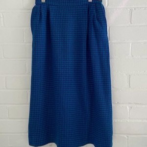 May include: A vintage blue houndstooth skirt hanging on a clear hanger. The skirt has a high waist and a classic A-line silhouette. The fabric is a woven material with a small check pattern in shades of blue and teal.