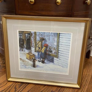 May include: Framed print of a winter scene with two children looking into a shop window. The artwork features a dog, a figure in a long coat, and a shop with the word "APHTA" visible. The frame is gold-colored.