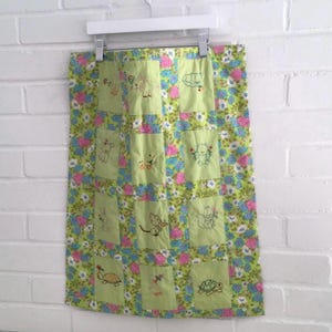 May include: A handmade baby quilt with a patchwork design. The quilt features embroidered animal designs on light green squares, surrounded by a floral patterned fabric in shades of green, pink, and blue. The quilt is hanging on a white hanger.