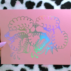 Peut inclure: Une carte rose avec une illustration holographique de deux chiens ressemblant à des caniches qui grognent. Les dents des chiens sont découvertes et le motif a un éclat métallique irisé. La carte est tenue sur un fond à imprimé léopard noir et blanc.