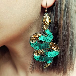 Puede incluir: Un par de pendientes de serpiente verde y dorado. Los pendientes están hechos de lentejuelas y cuentas y tienen forma de serpiente.