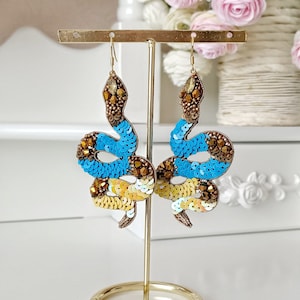 Puede incluir: Un par de pendientes de serpiente dorados con lentejuelas azules y amarillas. Los pendientes están en un soporte dorado.