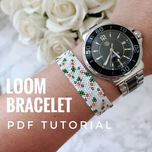 May include: A white and green loom bracelet with pink flowers. The bracelet is worn on a person's wrist with a silver watch.