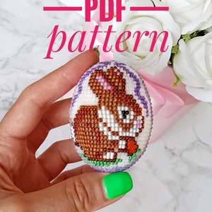 May include: A beaded Easter egg ornament featuring a brown bunny holding a carrot. The bunny is surrounded by purple, white and green beads. The egg is held in a hand with green nail polish. The text "PDF pattern" is written above the egg.