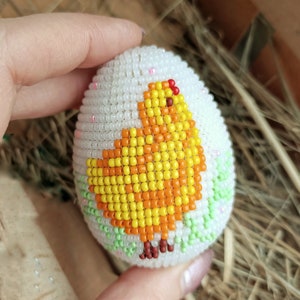 以下が含まれることがあります： 黄色とオレンジのビーズでできたひよこのデザインが施された、白いビーズでできたイースターエッグです。