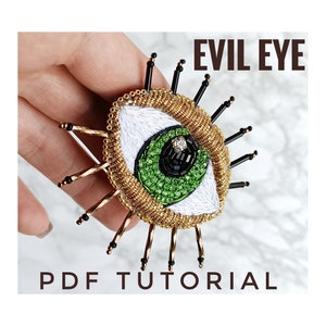 Puede incluir: Primer plano de un broche de ojo malvado dorado y verde con cuentas negras y un strass. La broche está siendo sostenida por una mano. El texto "EVIL EYE PDF TUTORIAL" está impreso debajo del broche.