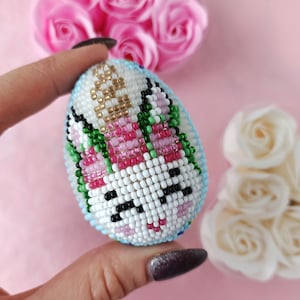 May include: A beaded Easter egg with a unicorn design. The egg is white with pink, green, and gold beads. The unicorn has a pink mane and tail, and a black nose and eyes.