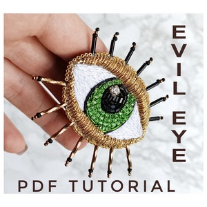 May include: A green and black evil eye brooch with gold beading and black accents. The brooch is held in a hand and the text "EVIL EYE PDF TUTORIAL" is visible in the background.